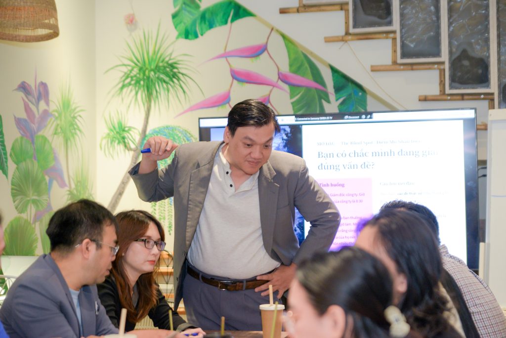 Nguyễn Chiến Trường đứng giữa phòng workshop, tay cầm bút, phía sau là màn hình chiếu câu hỏi "Bạn có chắc mình đang giải đúng vấn đề?".