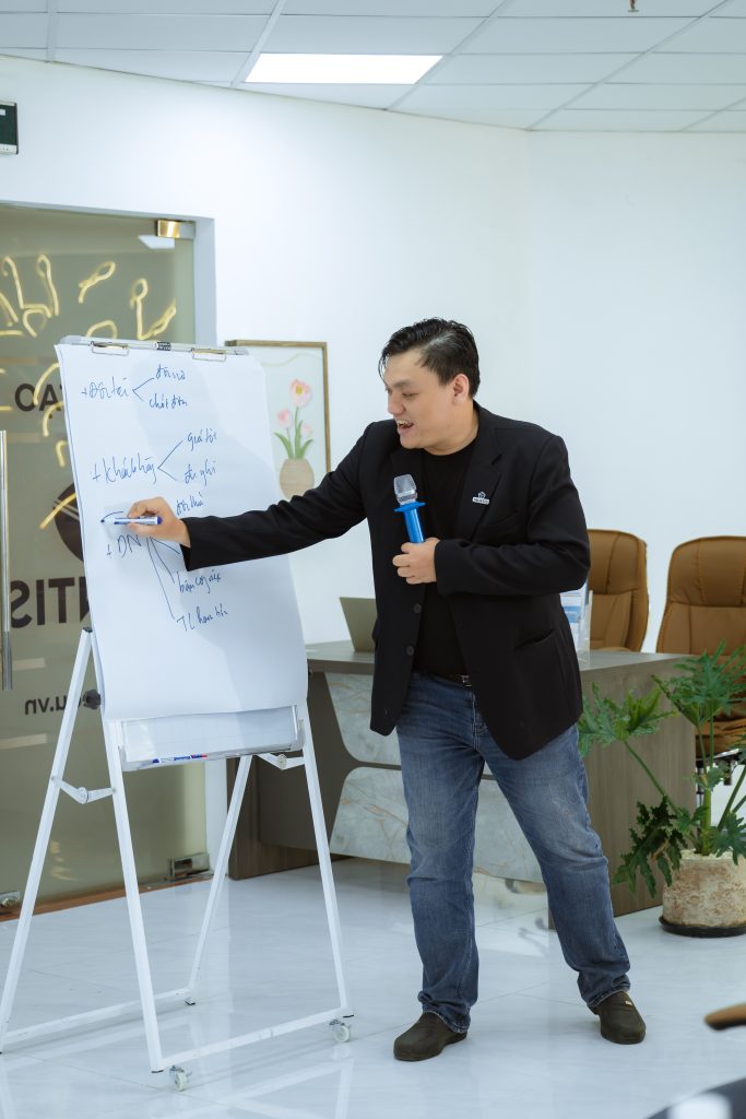 Nguyễn Chiến Trường cầm micro, đang viết lên bảng lật trong một văn phòng.