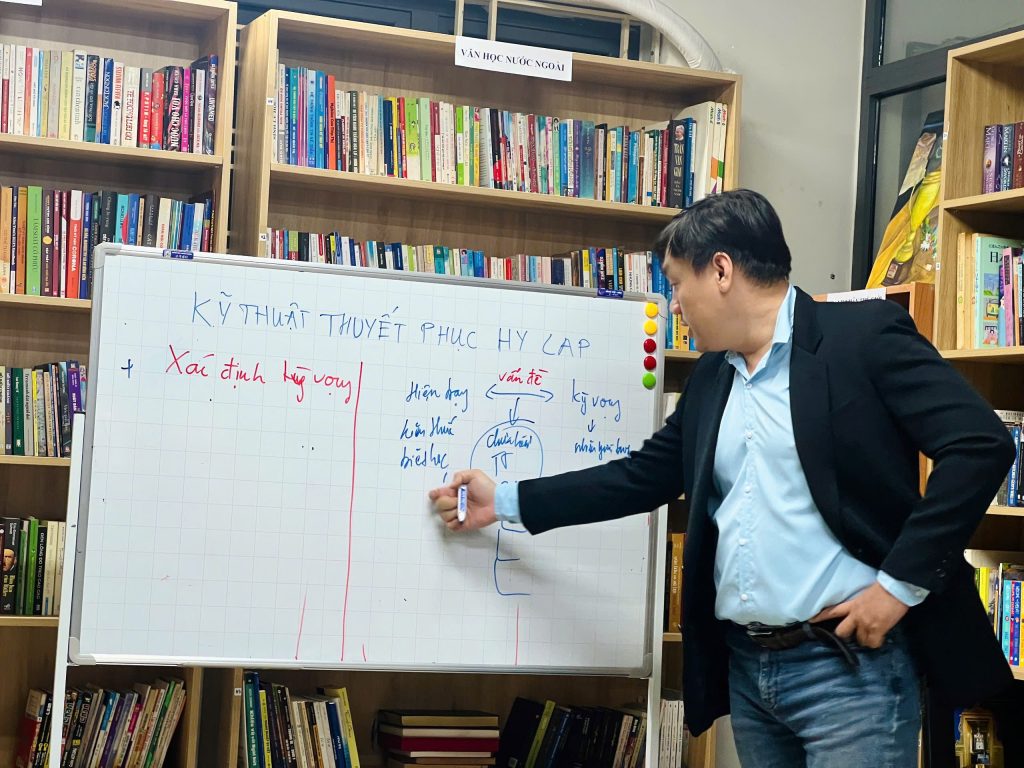 Nguyễn Chiến Trường đang viết lên bảng trắng ghi "Kỹ thuật thuyết phục Hy Lạp", phía sau là giá sách.