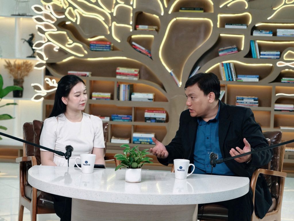 Nguyễn Chiến Trường trò chuyện với một người khách tại bàn tròn có micro, sau lưng là giá sách hình cây.